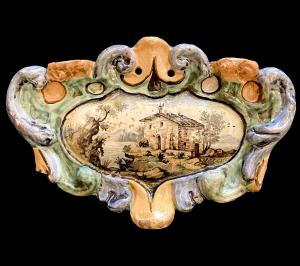 Formella in maiolica con bordo in rilievo rocaille e medaglione ovale centrale con scena architettonica e paesaggistica.Manifattura Chigi,San Quirico d’Orcia (Siena).