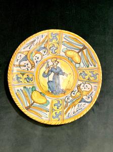 Piatto-tagliere in maiolica con decoro a quartieri a raffaellesche’motivi vegetali stilizzati e rocaille.Tondo centrale con figura di Santo.Manifattura di Deruta.