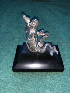 Figura de sereia prateada com cauda bifurcada segurando uma concha. Base em ébano.